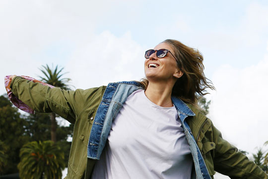 A white woman in sunglasses and a jacket runs, arms wide and smiling, in front of palm trees in San Francisco