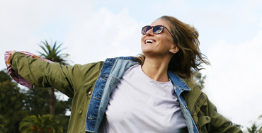 A white woman in sunglasses and a jacket runs, arms wide and smiling, in front of palm trees in San Francisco