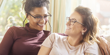 A young interracial lesbian couple, a black woman with braids and a white woman with tattoos, laugh together on their sofa