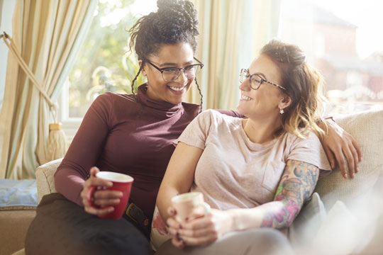 A young interracial lesbian couple, a black woman with braids and a white woman with tattoos, laugh together on their sofa