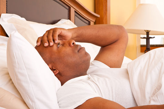 A black man in bed during the daytime. He has a flat expression with his hand to his head