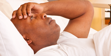 A black man in bed during the daytime. He has a flat expression with his hand to his head