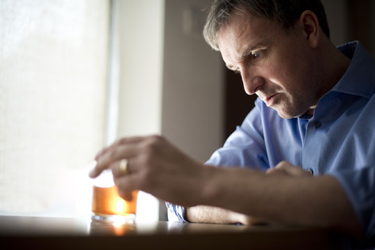 A middle-aged white man in a dress shirt, wearing a wedding ring, holds a glass of whiskey. He looks at it with a defeated expression.