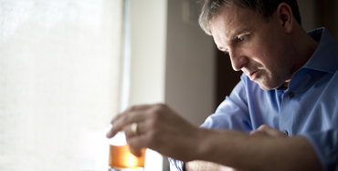 A middle-aged white man in a dress shirt, wearing a wedding ring, holds a glass of whiskey. He looks at it with a defeated expression.
